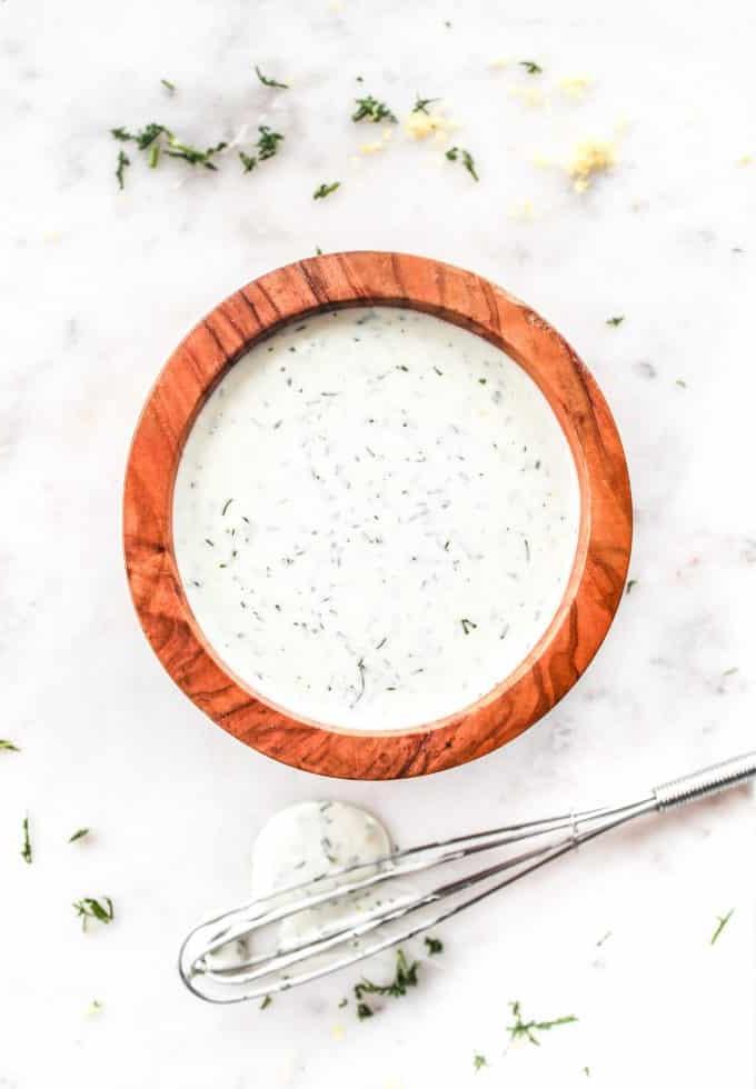 Einfache Zitronen-Dill-Soße 2 Eine kleine Glasschale gefüllt mit cremiger, gelblicher Zitronen-Dill-Soße, daneben frische Dillzweige und geschnittene Zitronen, auf einem hellen Holzbrett arrangiert.