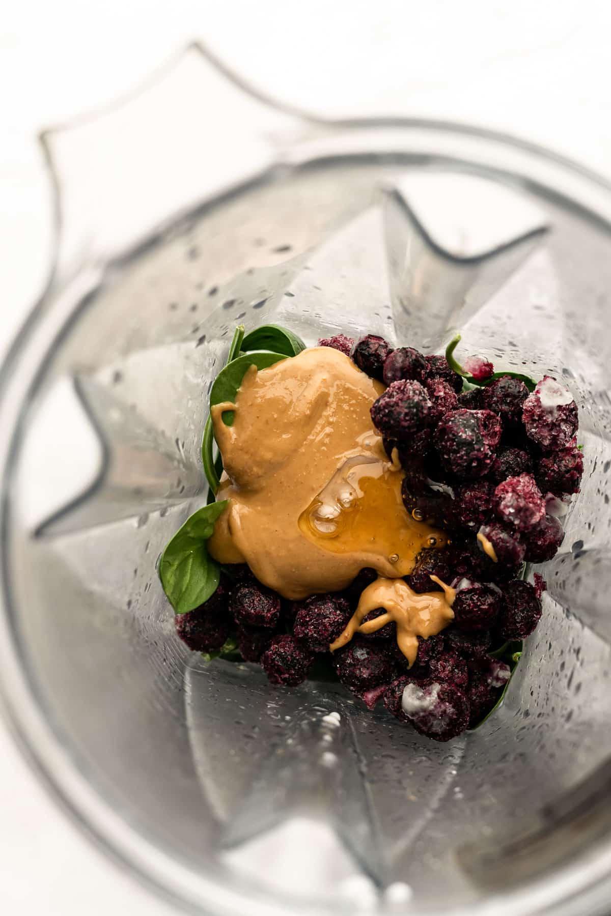Alle Hauptzutaten – gefrorene Blaubeeren, Spinat, Mandelbutter und Milch – bereit zum Pürieren in einem Hochleistungsmixer.