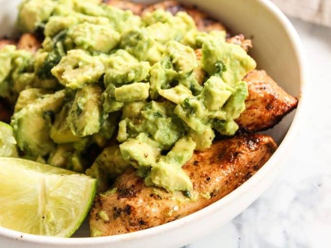 Ein einzelnes Stück gegrilltes Hähnchen mit stückigem Avocado-Topping auf einem weißen Teller.