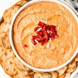 **Gerösteter Rote Paprika Hummus** 5 Nahaufnahme einer Schale mit Hummus, garniert mit frischen Kräutern und einem Löffel.