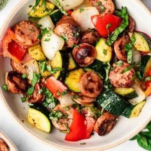 **20-Minuten-Wurst-Zucchini-Pfanne** 7 Die Autorin Christina, die eine Kochschürze trägt und lächelt.