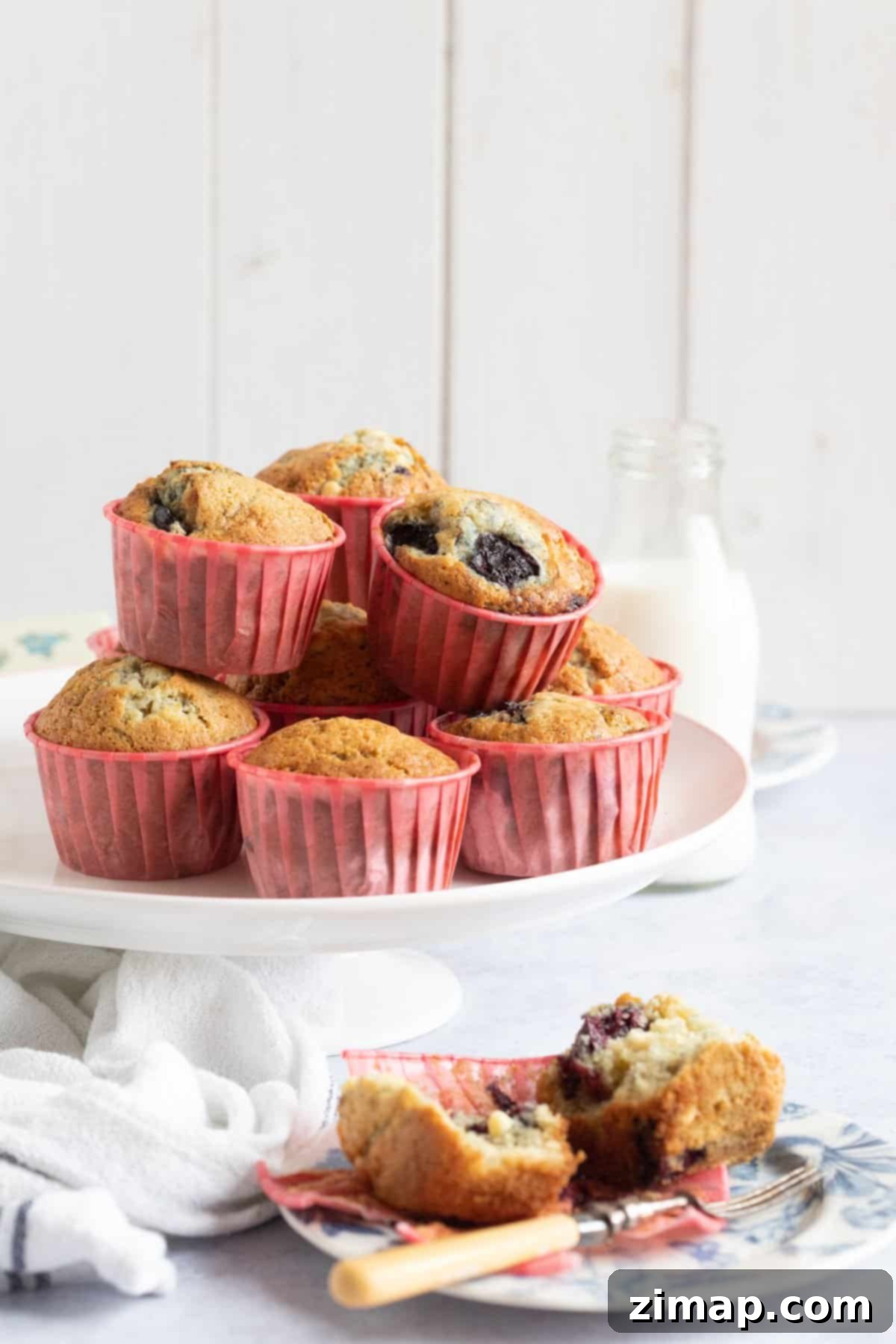 Εύκολα Μάφινς Λευκής Σοκολάτας με Κεράσια 3 Cherry muffins on a cake stand with one muffin on a blue plate in front of the cake stand.