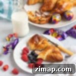 Απολαυστικά Κρουασάν με Αυγά Κρέμας στην Αερόφρυζα 9 Κρουασάν Creme Egg στη φριτέζα αέρος σε πιάτο με σμέουρα και βατόμουρα και ένα ποτήρι γάλα στο βάθος.