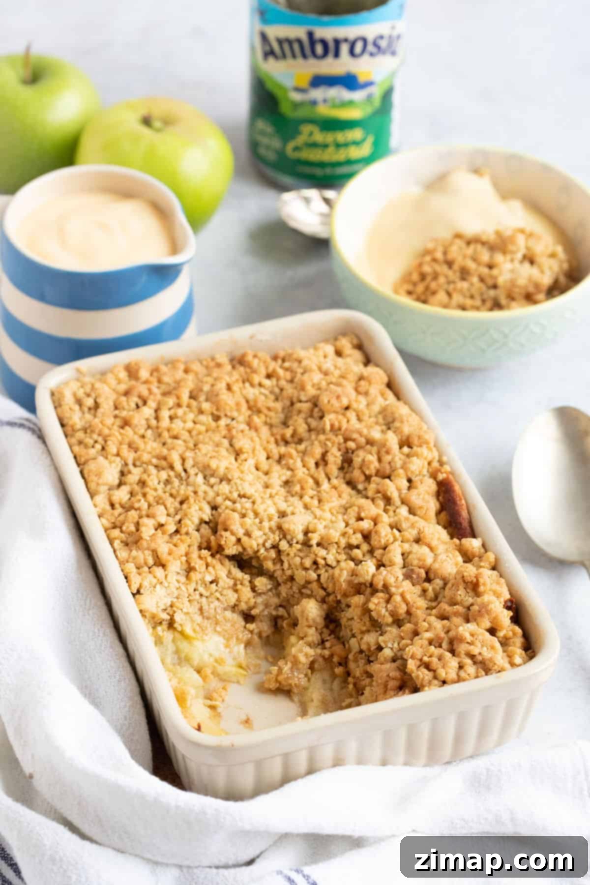 Εύκολο Κράμπλ Μήλου στο Air Fryer 2 Apple crumble σε φριτέζα αέρος σε ένα ταψί με μια κανάτα κρέμας κάσταρντ.