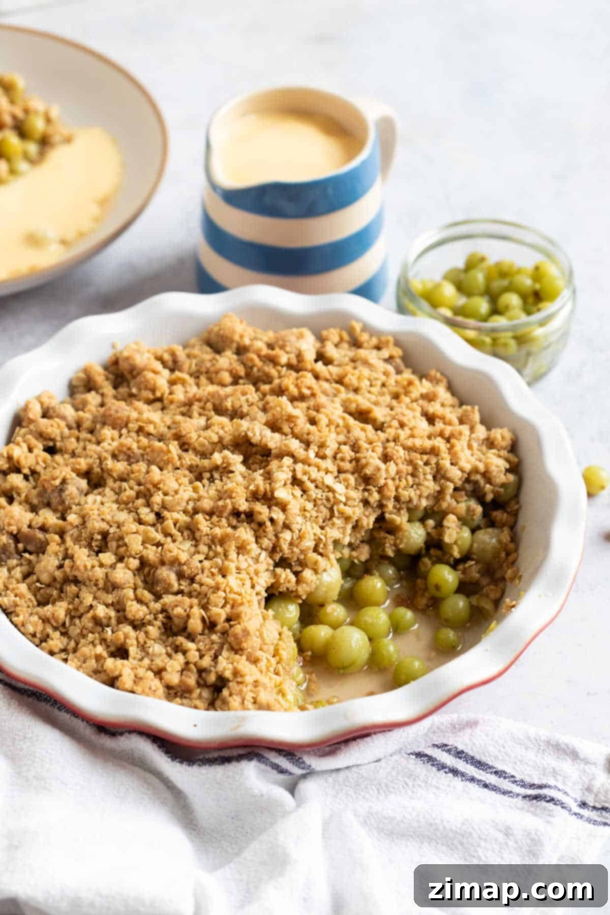 Κραμπλ Φραγκοστάφυλο χωρίς Κόπο 5 Crumble φραγκοστάφυλου σε κόκκινο και λευκό πιάτο πίτας με κανάτα κρέμας ζαχαροπλαστικής.