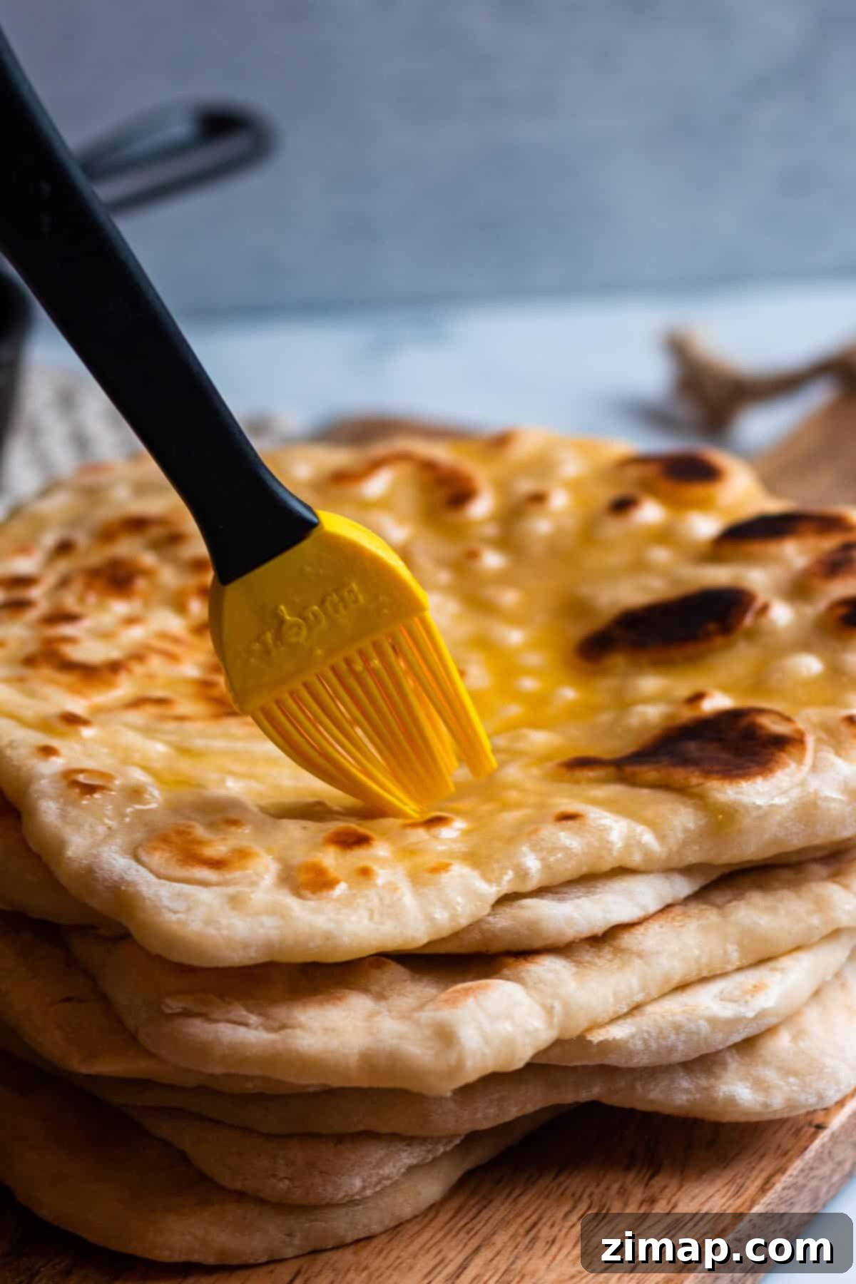 Προζυμένιο Νάαν με περίσσευμα 9 Μια στοίβα από φρεσκοψημένο sourdough naan με ένα πινέλο που απλώνει λιωμένο βούτυρο.