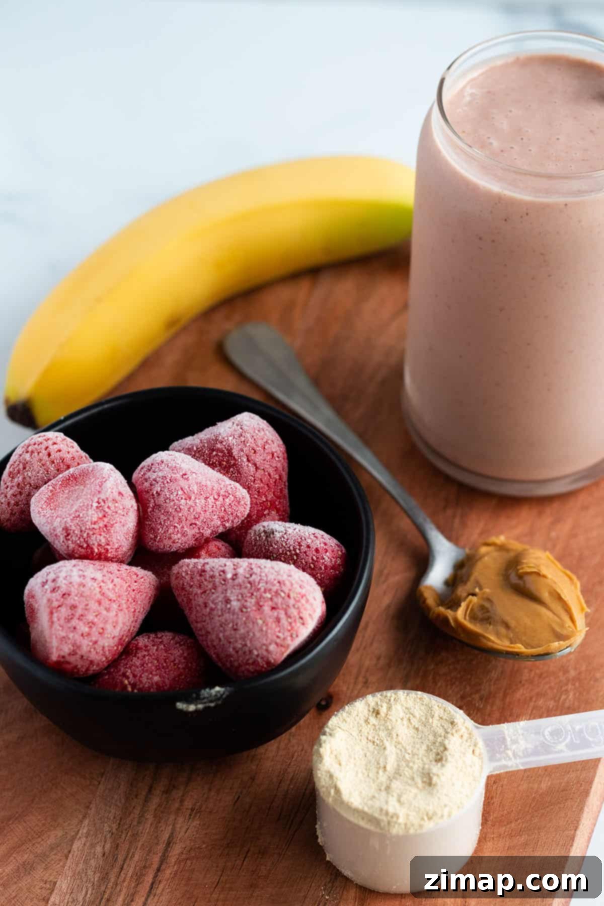 Πώς να φτιάξεις smoothie μπανάνας χωρίς γιαούρτι 2 Μια ξύλινη επιφάνεια με ένα smoothie, μπανάνα, κατεψυγμένες φράουλες, σκόνη πρωτεΐνης και φυστικοβούτυρο.