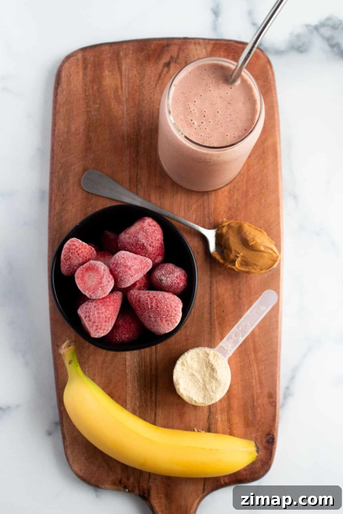 Πώς να φτιάξεις smoothie μπανάνας χωρίς γιαούρτι 6 Μια ξύλινη επιφάνεια με ένα ποτήρι γεμάτο smoothie, μπανάνα, μπολ με κατεψυγμένες φράουλες, φυστικοβούτυρο και σκόνη πρωτεΐνης.
