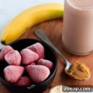 Πώς να φτιάξεις smoothie μπανάνας χωρίς γιαούρτι 7 Μια ξύλινη επιφάνεια με ένα ποτήρι γεμάτο ροζ smoothie, μπανάνα, κατεψυγμένες φράουλες, μια κουταλιά φυστικοβούτυρο και σκόνη πρωτεΐνης.