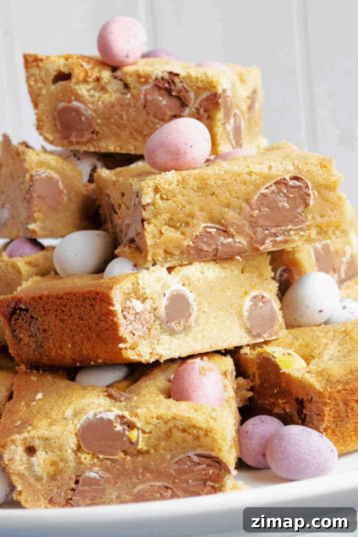 Εύκολες Μπάρες Μπισκότων Cadbury Μίνι Αυγά 7 Mini egg cookie bars stacked on a cake stand with extra mini eggs for decoration.