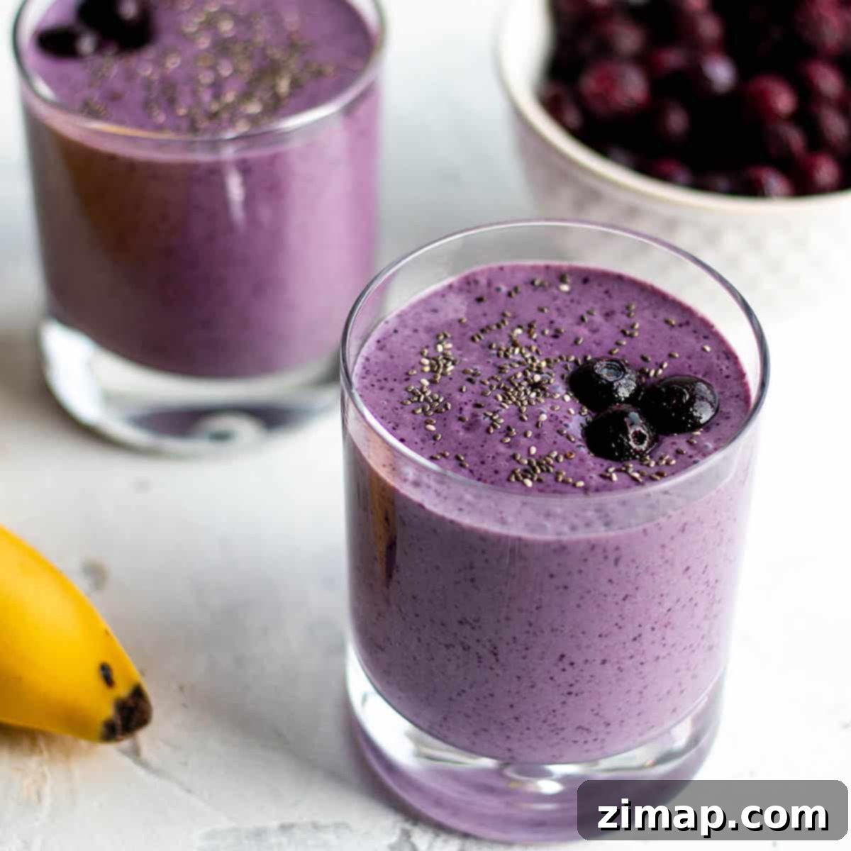Δύο μικρά ποτήρια με ένα μωβ smoothie, γαρνιρισμένο με μπλούμπερι, με ένα μπολ με μπλούμπερι και μια μπανάνα στο φόντο.